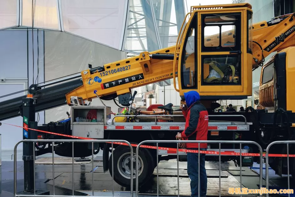 A construction worker wearing a blue hood stands next to a yellow crane truck at a worksite.