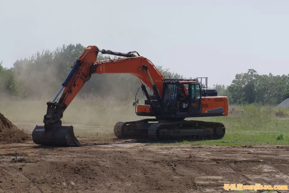An excavator levels dirt at an outdoor construction site with dust and trees visible.